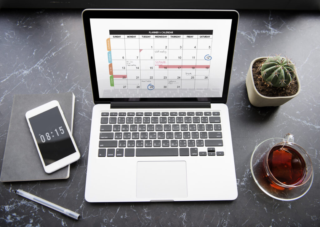 Computador em cima da mesa com calendário aparecendo na tela, um celular na mesa e uma xícara de café ao lado de uma flor.