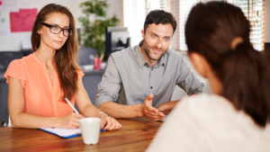 Reunião com dois contadores conversando com uma cliente em uma mesa de escritório. Um dos contadores está gesticulando, enquanto a outra faz anotações, ambos em um ambiente iluminado por luz natural.