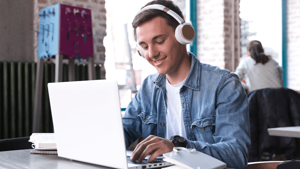 Jovem da geração Z sorridente usando fones de ouvido grandes e trabalhando em um laptop. Ele veste uma jaqueta jeans e está sentado em uma mesa, digitando no teclado, com um caderno ao lado. 