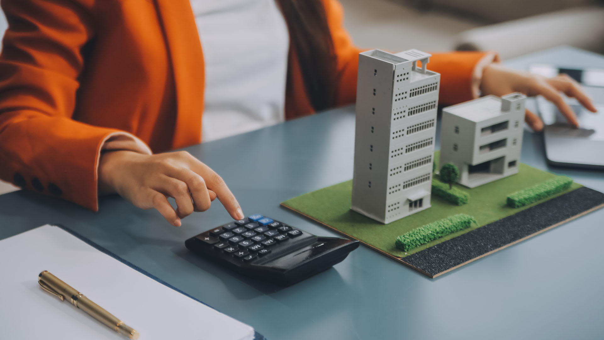 Pessoa usando uma calculadora em uma mesa, com maquetes de edifícios ao lado, sugerindo que ela está realizando a contabilidade do condomínio. Ela está vestida com um blazer laranja, e ao fundo há um notebook aberto, indicando uma possível atividade contábil.