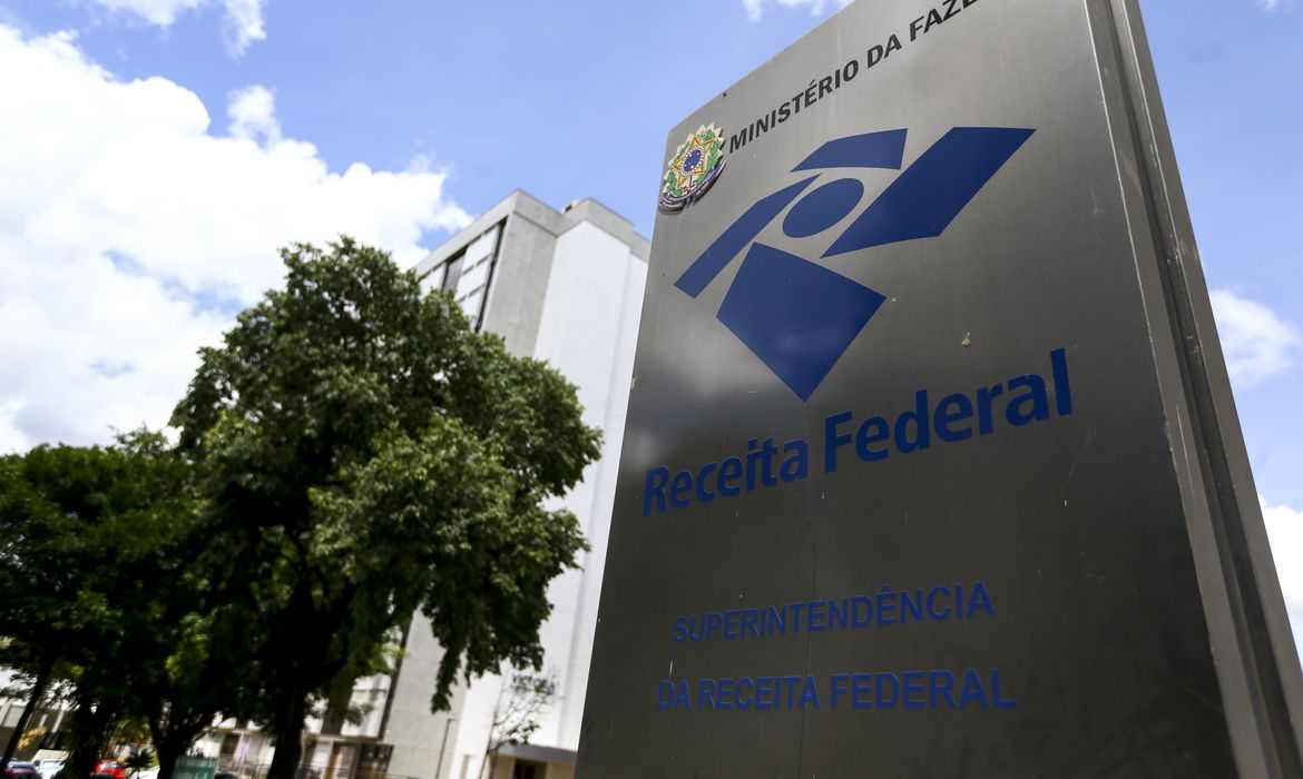 Placa da Receita Federal do Brasil, responsável pela mudança do CNPJ alfanumérico, em destaque, com o logotipo da instituição e os dizeres 'Superintendência da Receita Federal' abaixo. Ao fundo, uma árvore e um edifício de múltiplos andares são parcialmente visíveis sob um céu azul com algumas nuvens claras. 
Imagem por Marcelo Camargo da Agência Brasil.