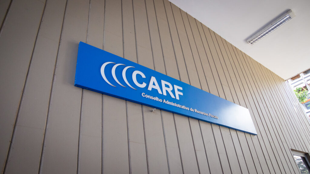 Mesa de julgamento do CARF diante de painel digital com dados de receitas financeiras e tributos PIS COFINS